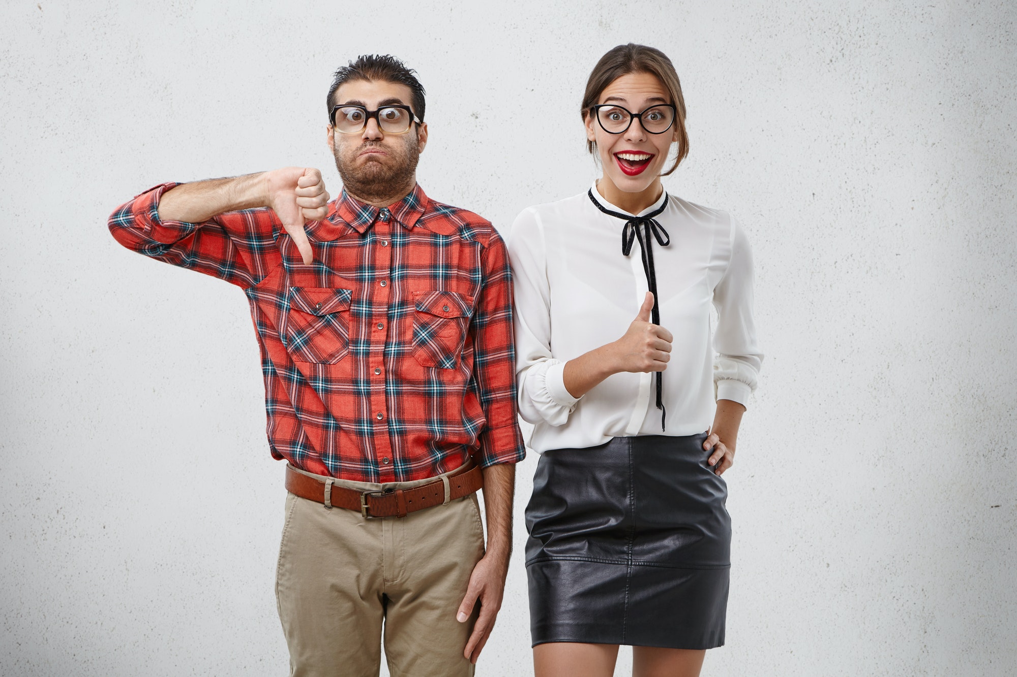 Displeased funny nerd shows thumb down, has sulk expression, stands next to happy female model who e