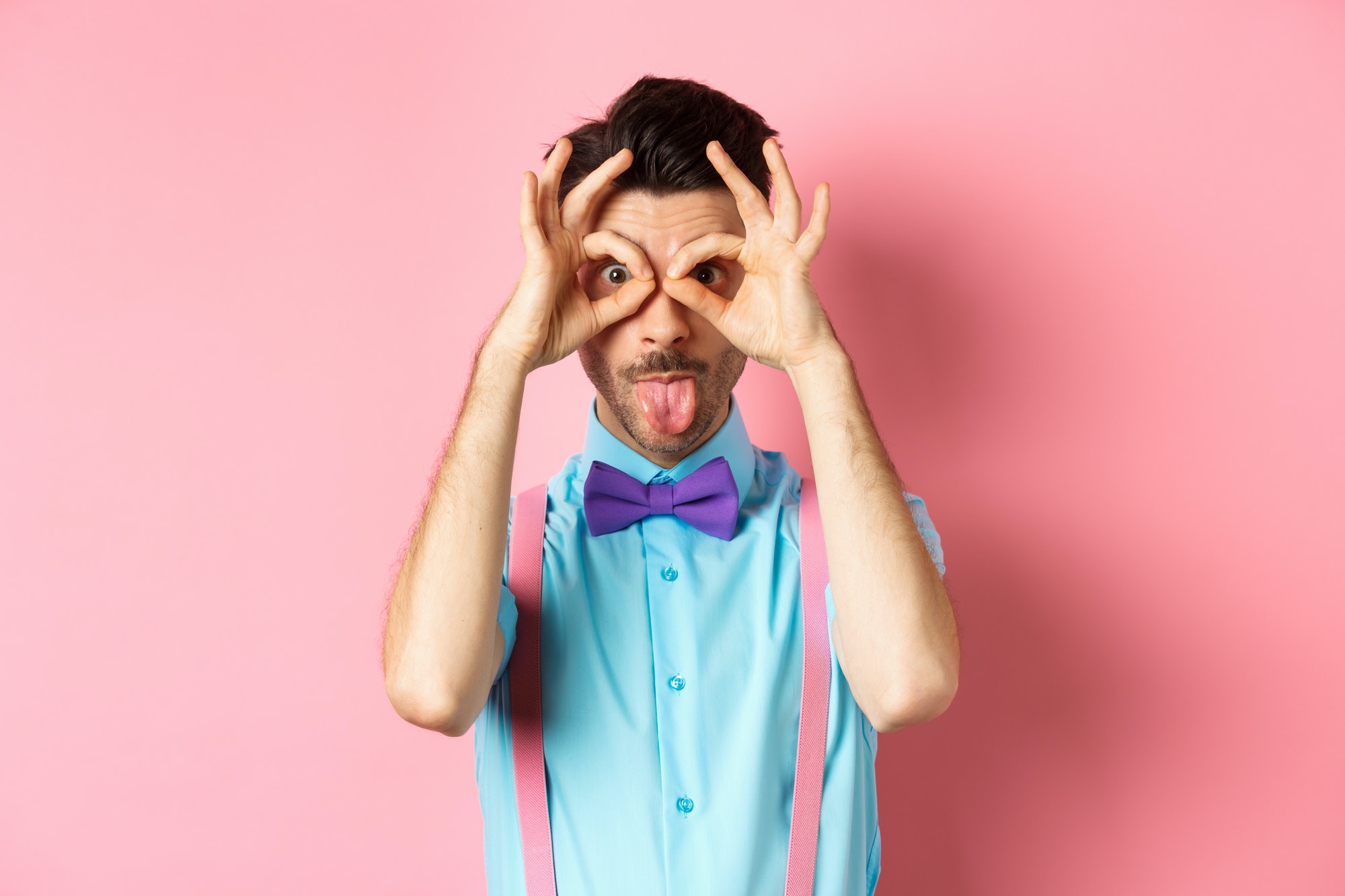 Funny and silly young man in bow-tie and suspenders, looking through hand-binoculars and showing