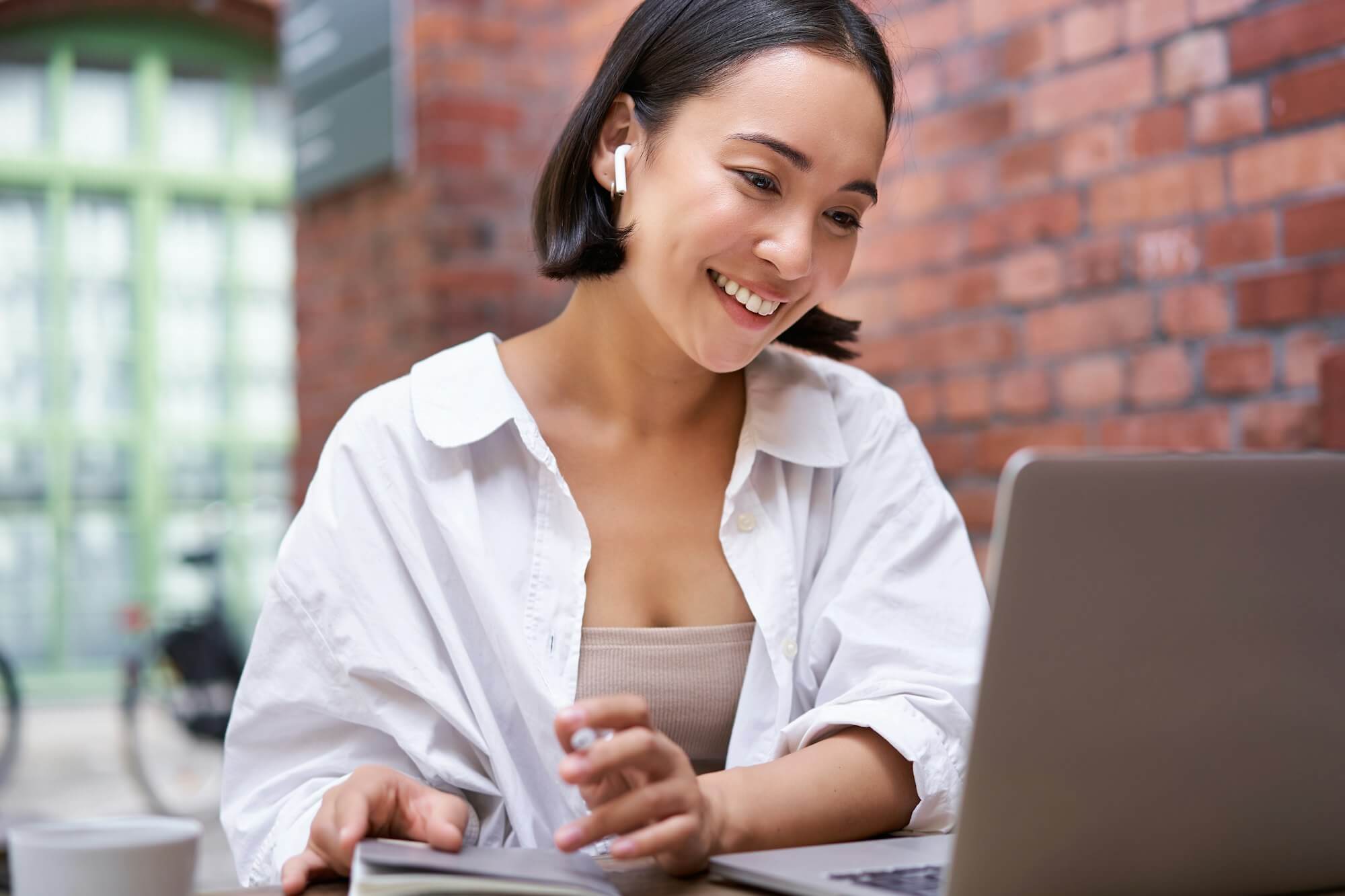 Young smiling asian woman with wireless earphones, sitting in coworking office space with laptop